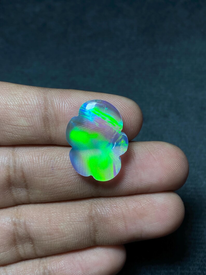 May include: A colorful, iridescent opal gemstone with a unique, flower-like shape. The stone displays vibrant green, blue, and purple hues, with a polished surface. The gemstone is held in a hand, showcasing its natural beauty.