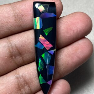 Op de afbeelding: Een lange, taps toelopende zwarte hanger met een inleg van iriserende, veelkleurige geometrische scherven. Het oppervlak is glad en gepolijst en vertoont levendige flitsen van groen, roze, blauw en geel licht op het donkere basismateriaal.