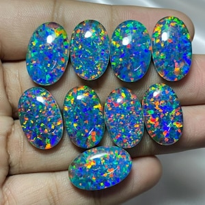May include: A collection of nine oval-shaped opals. The opals display a dark blue base color with vibrant flashes of green, orange, and yellow. The gemstones are held in a hand, showcasing their polished surfaces and iridescent play-of-color.
