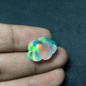 May include: A cloud-shaped gemstone with iridescent colors of green, blue, and orange. The gemstone is held in a hand, showcasing its unique shape and vibrant play of light. The stone appears to be polished and smooth.