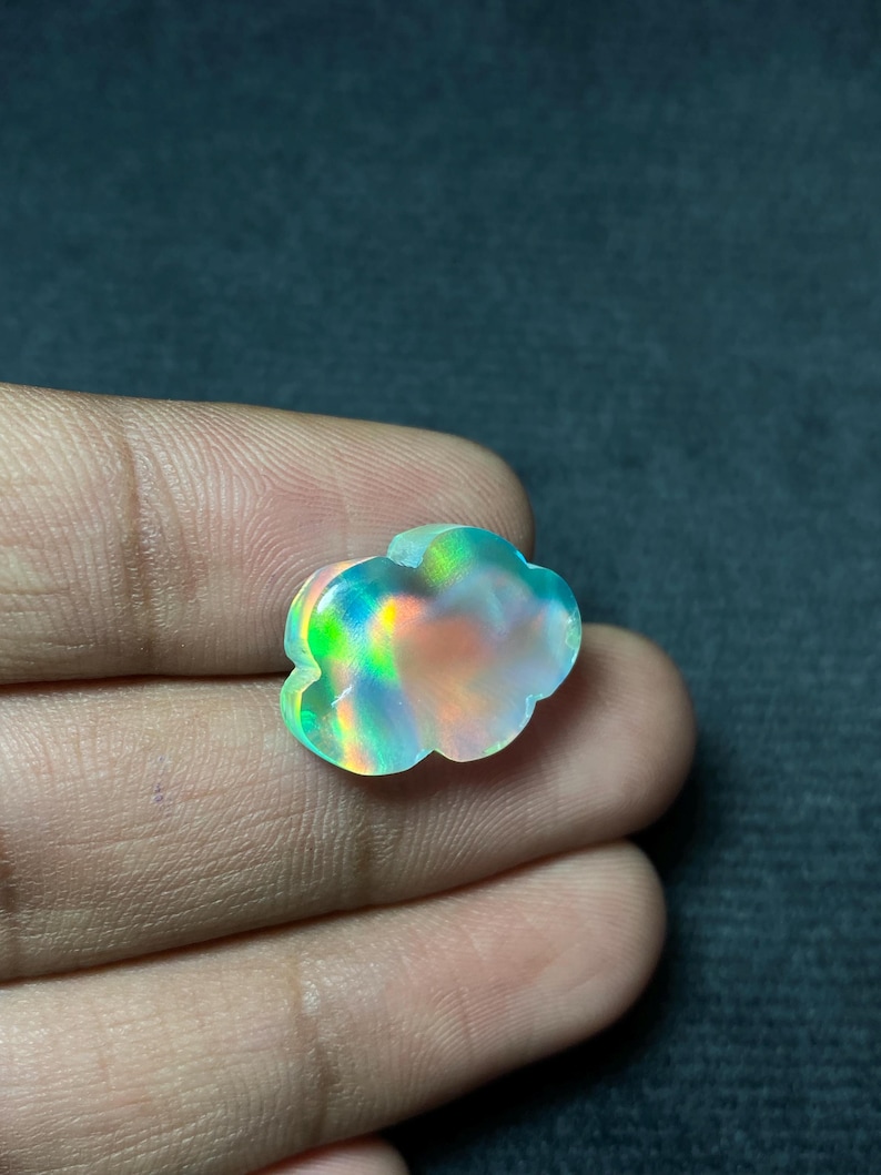 May include: A cloud-shaped gemstone with iridescent colors of green, blue, and orange. The gemstone is held in a hand, showcasing its unique shape and vibrant play of light. The stone appears to be polished and smooth.