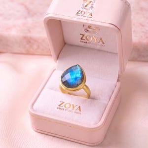 May include: A gold ring featuring a pear-shaped, faceted blue labradorite gemstone, displayed inside a light pink velvet-lined jewelry box. The box lid and interior are printed with the Zoya Gems Store India logo in gold lettering.