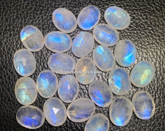 May include: A collection of oval-shaped moonstone cabochons, with a milky white color and iridescent blue and green flashes. The stones are arranged in a loose pile on a black surface.