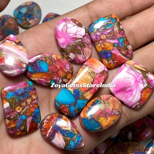 Könnte beinhalten: Sortiment an bunten, polierten und gemusterten Edelsteinkabochons. Die Cabochons zeigen eine Mischung aus rosa, orange, türkis und goldenen Farben. ZoyaGemsStoreIndia ist auf dem Bild gedruckt.