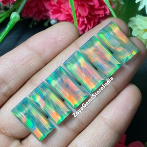 May include: Five rectangular-shaped, iridescent opal gemstones with a green, blue, and orange color scheme. The gemstones are arranged in a row, with the largest gemstone at the top and the smallest gemstone at the bottom. The gemstones are being held in a hand.