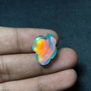 May include: A colorful, iridescent gemstone with a unique, flower-like shape. The stone displays vibrant hues of orange, blue, and green, creating a striking visual effect. The gemstone is held in a hand against a dark background.