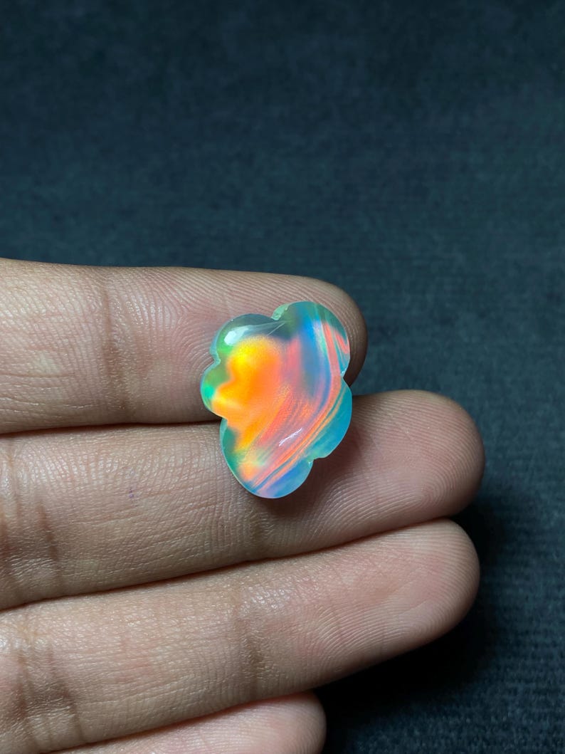 May include: A colorful, iridescent gemstone with a unique, flower-like shape. The stone displays vibrant hues of orange, blue, and green, creating a striking visual effect. The gemstone is held in a hand against a dark background.