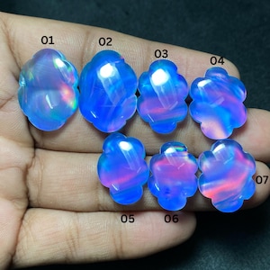 May include: Seven iridescent, flower-shaped gemstones in shades of blue, pink, and purple are displayed. Each stone has a unique pattern and internal glow. The gemstones are held in a hand, showcasing their color variations and shapes.