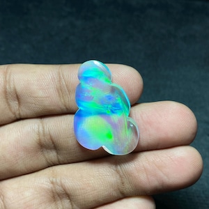 May include: A polished, iridescent opal gemstone with a unique, organic shape. The opal displays a vibrant play of color, including blues, greens, and hints of yellow. The gemstone is held in a hand, showcasing its size and luster.