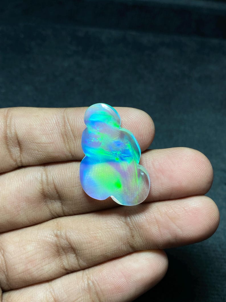 May include: A polished, iridescent opal gemstone with a unique, organic shape. The opal displays a vibrant play of color, including blues, greens, and hints of yellow. The gemstone is held in a hand, showcasing its size and luster.
