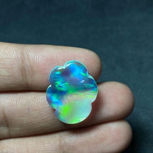 May include: A colorful, iridescent gemstone with a unique, flower-like shape. The stone displays a vibrant array of blues, greens, and purples, with a pearly, opalescent sheen. The gemstone is held in a hand.