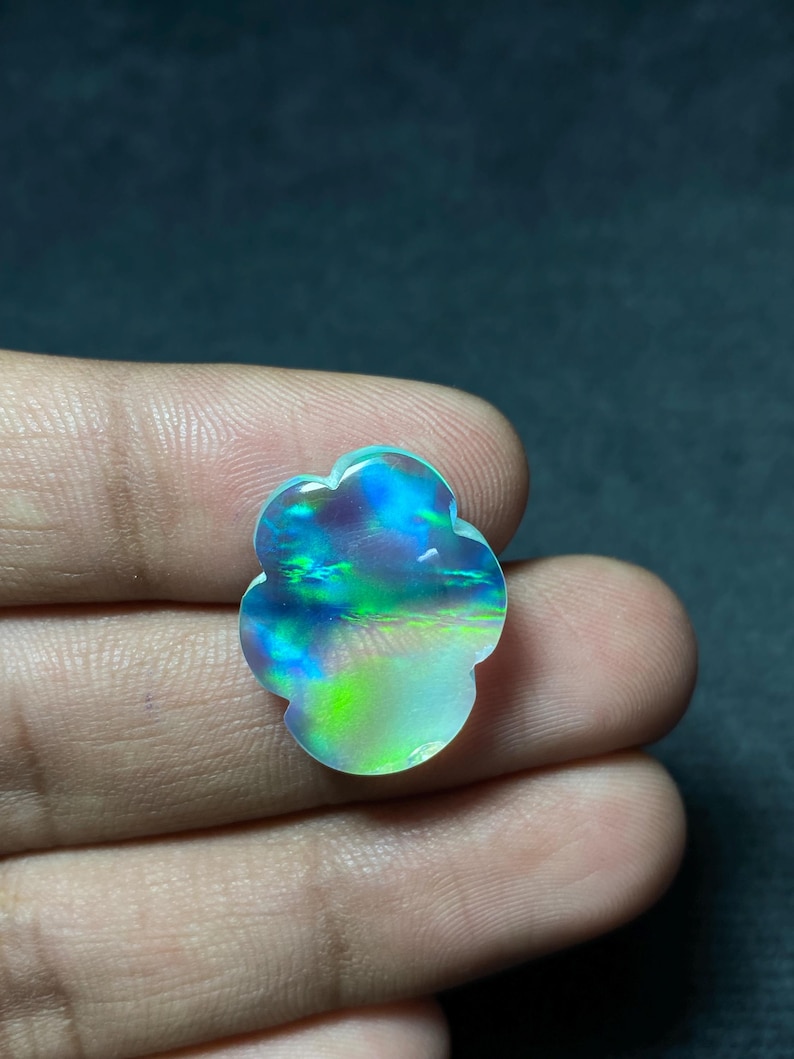 May include: A colorful, iridescent gemstone with a unique, flower-like shape. The stone displays a vibrant array of blues, greens, and purples, with a pearly, opalescent sheen. The gemstone is held in a hand.