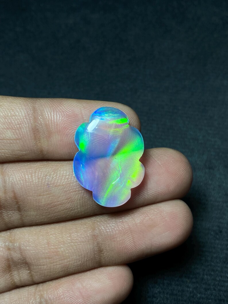 May include: A polished, iridescent opal gemstone in a flower shape. The opal displays a vibrant play of color, with hues of blue, green, and purple. The gemstone is held in a hand against a dark background.