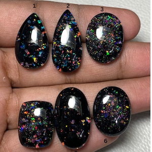 Puede incluir: Seis piedras preciosas de ópalo negro en varias formas, incluyendo lágrima y ovalada. Cada piedra muestra un vibrante juego de colores con destellos de rojo, verde, azul y amarillo. Los ópalos están pulidos y tienen un acabado brillante.