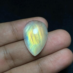 May include: A pear-shaped, iridescent gemstone with a polished surface. The stone displays a play of colors, including yellow, blue, and green, against a translucent background. The gemstone is held in a hand against a dark background.