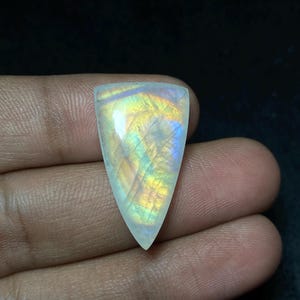 以下が含まれることがあります： 三角形の形をした、研磨された虹色のムーンストーンの宝石。石は、青、黄、緑のきらめきを伴う色の遊びを示しています。宝石は暗い背景に対して手で保持されています。