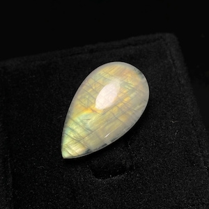 以下が含まれることがあります： 磨かれた表面を持つ、水滴型の虹色の宝石。石は、黄色、緑、白などの色の混合を示し、微妙な内部の輝きがあります。宝石は黒い背景に配置されています。