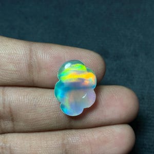 May include: A cloud-shaped opal gemstone with a vibrant display of colors, including green, yellow, and blue. The gemstone is held in a hand, showcasing its iridescent qualities. The opal's unique shape and color make it a striking piece of jewelry.