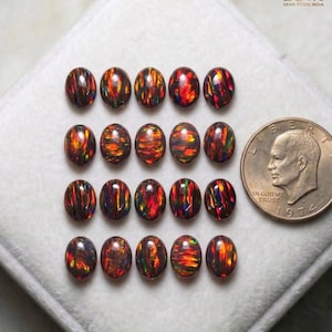 Puede incluir: Dieciséis piedras preciosas de ópalo negro de forma ovalada con destellos rojos, naranjas y verdes. Los ópalos están dispuestos sobre una superficie blanca junto a una moneda de veinticinco centavos estadounidense. Las piedras preciosas son de Zoya Gems Store India.