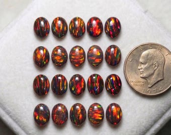May include: Sixteen oval-shaped black opal gemstones with vibrant red, orange, and green flashes. The opals are arranged on a white surface next to a US quarter coin. The gemstones are from Zoya Gems Store India.