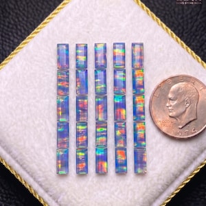 May include: Twenty-five rectangular baguette-cut opals arranged in five vertical rows of five. Each gemstone displays a translucent blue base with vibrant, multi-colored iridescent flashes of red, green, and yellow. A 1974 U.S. penny is shown for scale.