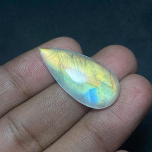 May include: A pear-shaped, iridescent moonstone gemstone. The polished stone displays a mix of colors, including yellow, blue, and white, with internal lines and a translucent appearance. The gemstone is held in a hand against a dark background.
