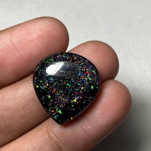 以下が含まれることがあります： 鮮やかな多色インクルージョンを持つ、ハート型のブラックオパール宝石。研磨された表面は光を反射し、緑、赤、青などの色のスペクトルを示しています。宝石は手で持たれており、そのサイズと形状が強調されています。