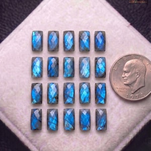 Pode incluir: Dezasseis pedras preciosas retangulares de labradorite com superfícies facetadas, exibindo tons iridescentes azuis e verde-água. Uma moeda de um quarto de dólar dos EUA está ao lado das pedras preciosas. As pedras preciosas estão dispostas sobre uma superfície branca. O logotipo Zoya Gems Store India está no canto superior direito.