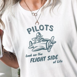 May include: White t-shirt with a blue graphic of a pilot flying a plane. The text "PILOTS Look on the FLIGHT SIDE of Life" is printed on the shirt.