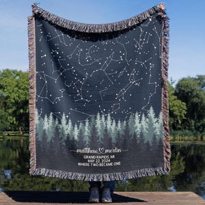 May include: A dark blue woven blanket featuring a constellation design with white connecting lines and stars. The bottom edge displays a forest silhouette in shades of green and gray. The blanket has a fringed edge and includes the text "matthew ♡ merlin", "GRAND RAPIDS, MI MAY 22, 2024 WHERE TWO BECAME ONE".
