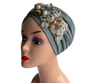 May include: A gray turban with a decorative floral embellishment featuring gold and white accents. The embellishment includes pearls, rhinestones, and fabric flowers.