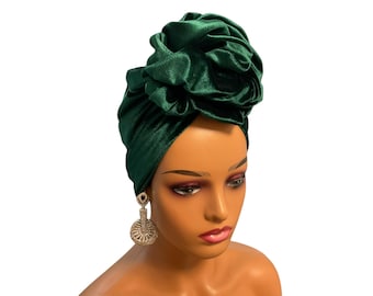 May include: A dark green velvet turban featuring a large, gathered fabric flower detail on the top. The headpiece is displayed on a mannequin head, which is also wearing a pair of dangling, circular, gold-toned earrings with clear crystals.