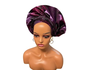 May include: A mannequin head wearing a structured, metallic purple headwrap with a dark floral pattern. The headwrap features intricate folds and a high, rounded shape. The mannequin has light brown skin, dark makeup, and gold hoop earrings.