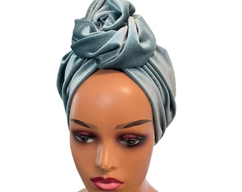 May include: A light blue velvet turban featuring a large, knotted fabric rose detail at the top center. The head covering is crafted from a soft, plush material with a subtle sheen, designed to wrap securely around the head with draped folds.