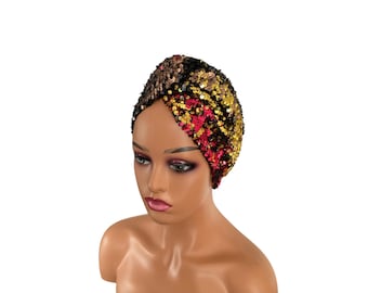 May include: A sequined turban in a mix of colors including black, gold, and red. The head covering is displayed on a mannequin head with a neutral skin tone. The sequins create a textured, shimmering effect.