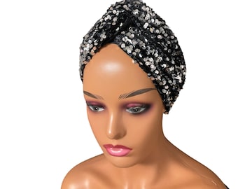 May include: A black turban-style head wrap covered in silver and black sequins. The head covering is on a mannequin head with dark eye makeup and pink lipstick. The background is white.