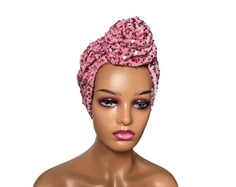 May include: A pink sequined turban head wrap featuring a decorative knot detail at the front. The fabric is densely covered in small, shimmering pink sequins that create a textured, reflective surface across the entire head covering.