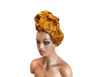 May include: A mustard gold headwrap featuring a textured, crinkled fabric surface styled into large, voluminous rosette-like folds. The headwrap is displayed on a mannequin head, which is also wearing a pair of gold-toned circular drop earrings.