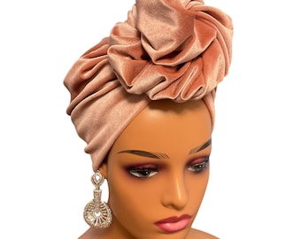 May include: A dusty rose velvet turban with a large, gathered knot on top. The turban is worn on a mannequin with a neutral makeup look.
