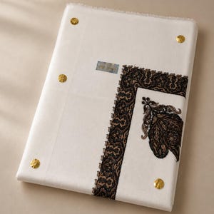 May include: A rectangular white fabric textile featuring a dark brown embroidered border with intricate floral patterns. The fabric is accented with four small, circular gold-colored metallic studs placed near the corners and edges.