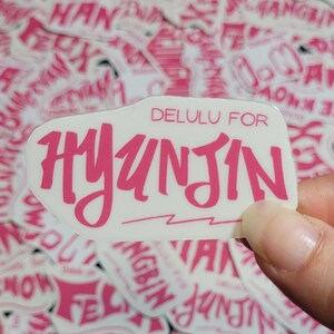 May include: White sticker with pink lettering that reads "DELULU FOR HYUNJIN" with a pink lightning bolt underneath. The sticker is slightly curved and has a glossy finish.
