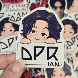 May include: A die-cut vinyl sticker featuring a cartoon illustration of a person with dark wavy hair resting their chin on a white rectangular sign. The sign displays the black text DPR IAN in a stylised font. The sticker has a white border.
