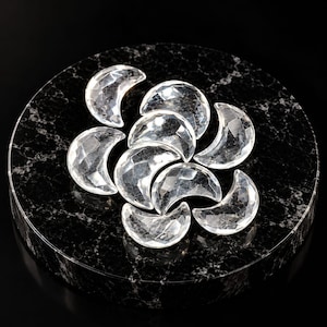 May include: A collection of nine clear, faceted crystal moon-shaped cabochons arranged in a cluster. Each piece features a crescent form with multiple geometric facets that reflect light, creating a sparkling, transparent appearance.