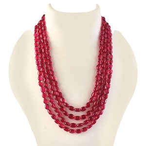Op de afbeelding: Een meerstrengs ketting met ovale, dieprode kralen. De ketting wordt getoond op een witte mannequin buste, die het gelaagde ontwerp en de kleur van de sieraden benadrukt. De kralen hebben een gepolijste afwerking.