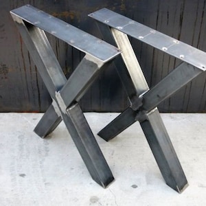 May include: Two black metal table legs in an X shape. The legs are made of thick, rectangular metal and are welded together at the corners. The legs are designed to support a table top.