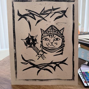 May include: A black ink block print on tan paper featuring a cat wearing a helmet and holding a spiked mace. The cat is centered between two horizontal rows of thorny branches, all enclosed within a rectangular border frame.