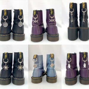 May include: A set of six black leather boots with silver charms attached to the laces. The charms are different designs, including a crescent moon, a sun, a spider, and a small figure. The boots are all the same style, with a lace-up front and a chunky heel.