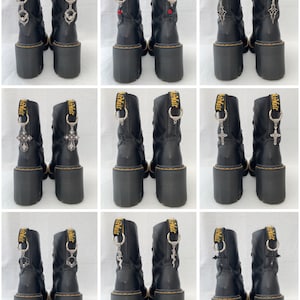 May include: Nine pairs of black leather boots with different silver charms attached to the laces. The charms include a cross, a bat, a heart, and a skull. The boots have a yellow AirWair label on the back.