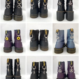 May include: Nine pairs of black leather boots with various charms attached to the laces. The charms include a heart, a cross, a sunflower, a crescent moon, a pentagram, and a butterfly. The boots are all the same style, with a black leather upper, yellow stitching detail, and a black rubber sole.
