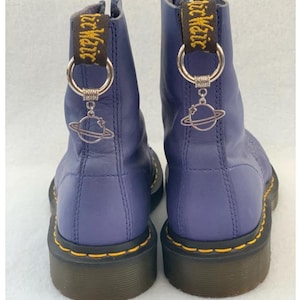 May include: A pair of purple leather Dr. Martens boots with yellow stitching and black soles. Each boot has a silver charm attached to a black strap that says "Dr. Martens". The charms are of a planet with a star.